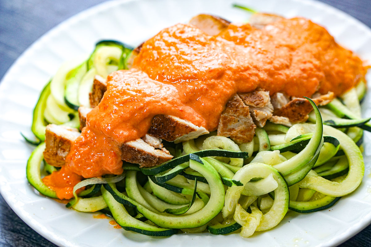 Closeup of a white plate with zucchini noodles and sliced cooked chicken breast topped with the finished sauce.
