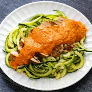 White plate with veggie noodles and chicken breast topped creamy red pepper sauce.