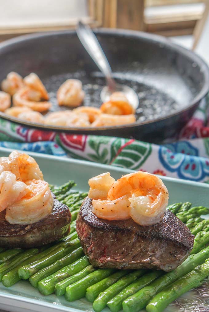 Long photo of steaks with a pan of shrimp in background.