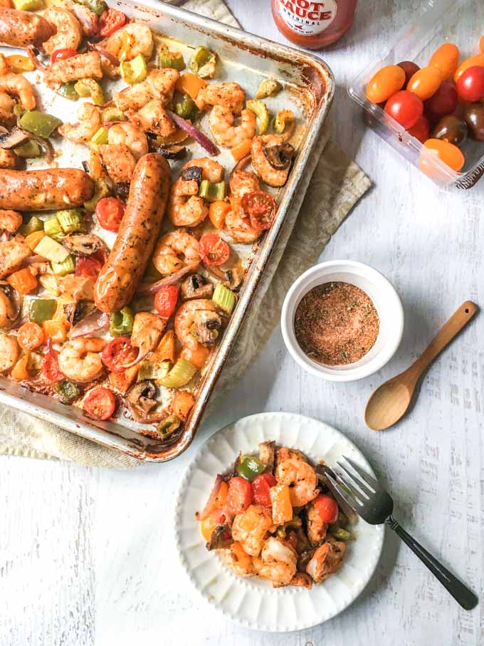 Long photo of sheet pan full of cajun chicken, shrimp, sausage and vegetables. and a small white plate with a serving on.