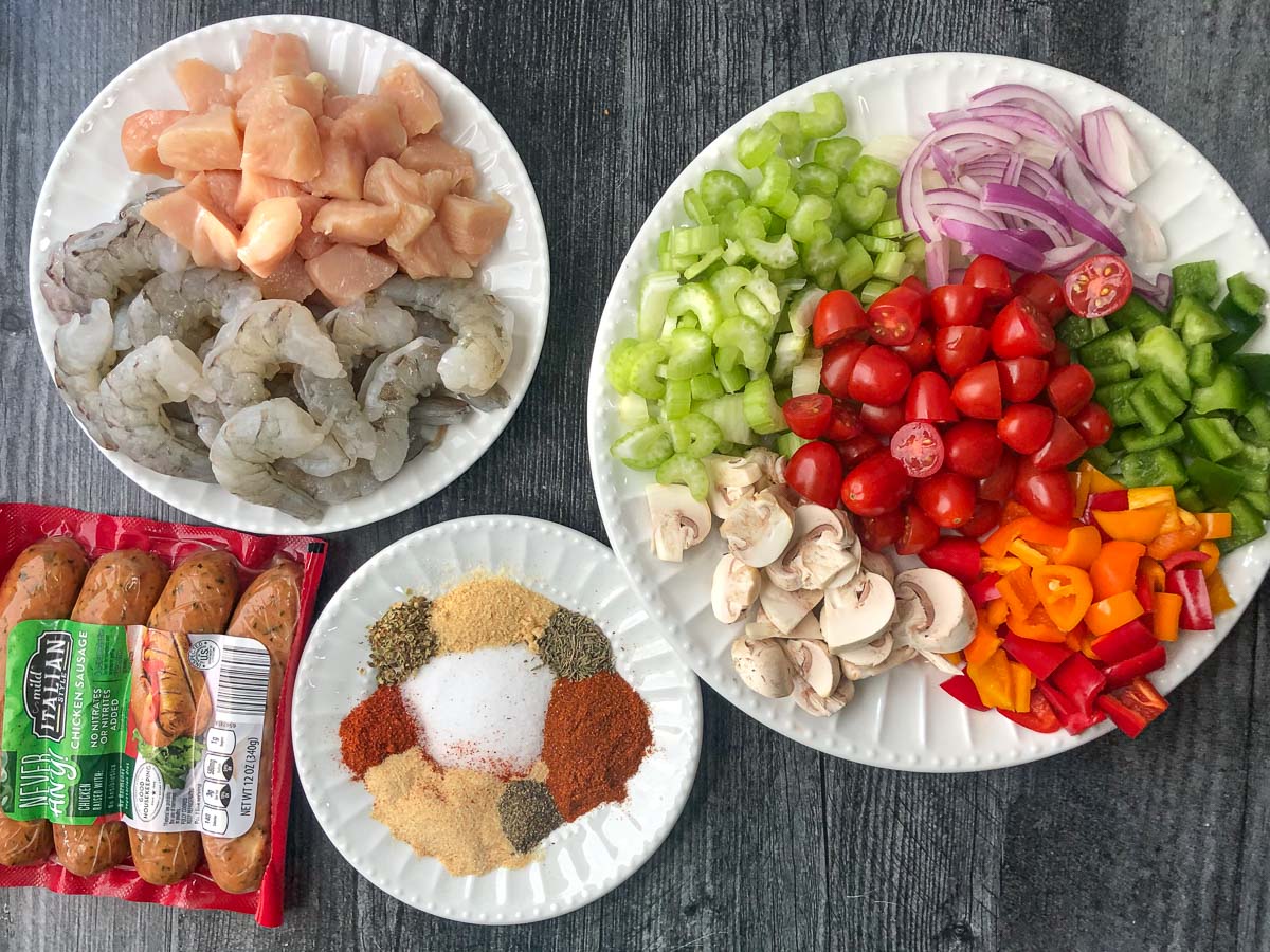 recipe ingredients - chopped veggies, raw shrimp and chicken, sausages and a plate of cajun spices