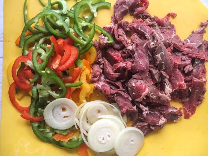 yellow cutting board with raw steak, onions and peppers
