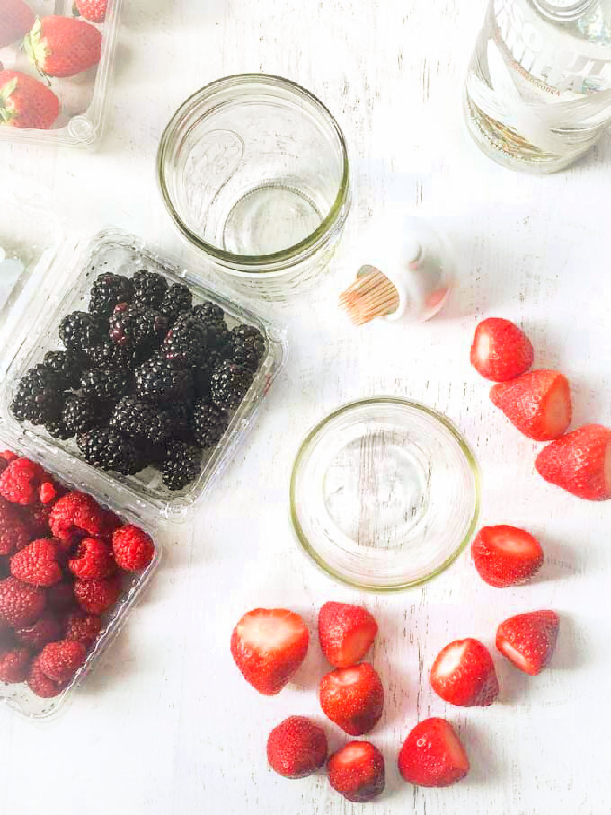 Aerial view of recipe ingredients of fresh strawberries, black berries, empty jars and a bottle of vanilla vodka.