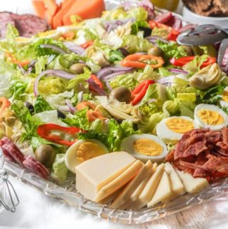 Large platter of salad, cheese and meats with a salt container and wooden spoon.