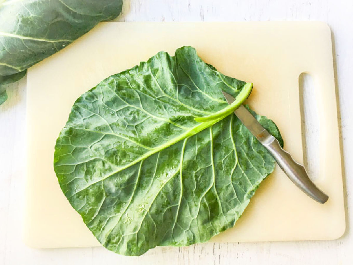 White cutting board with a large collard green leaf with a knife slicing the vein in the middle. 