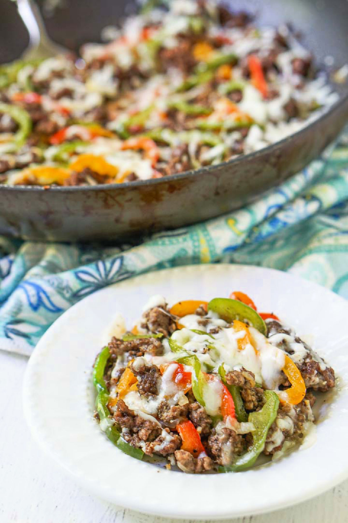 A white plate with a serving of hamburger meat and peppers covered with cheese and a pan in the background.