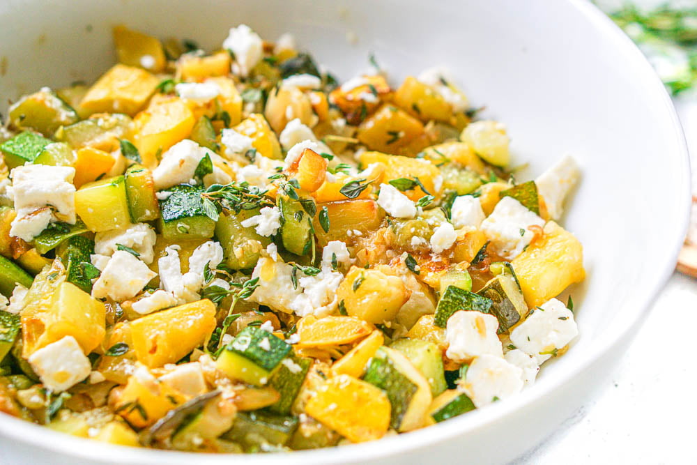 closeup of a white bowl filled with yellow zucchini and chunks of feta and fresh thyme leaves