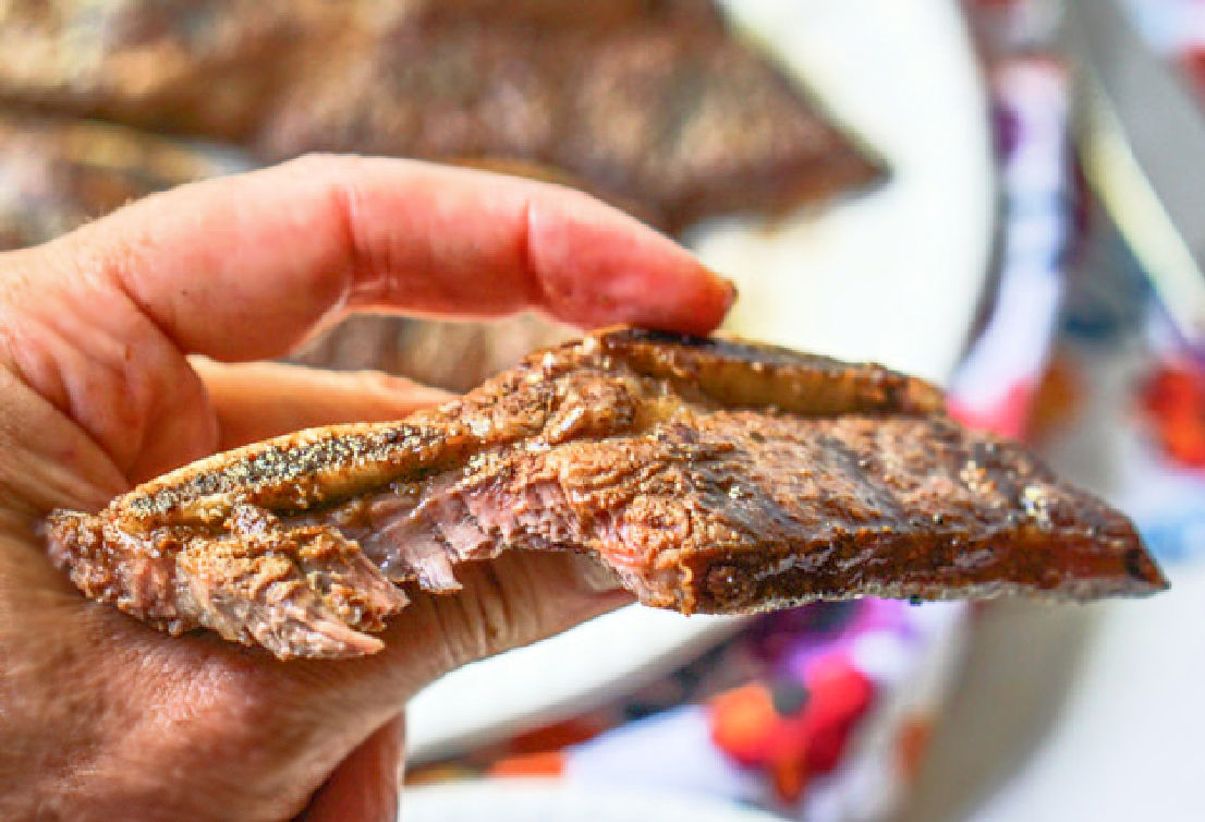 A hand holding a flanken beef rib with a bite taken out.