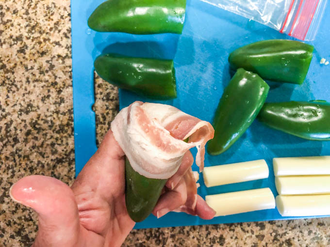 hand holding jalapeno wrapped with raw bacon and cutting board with raw peppers and cheese sticks