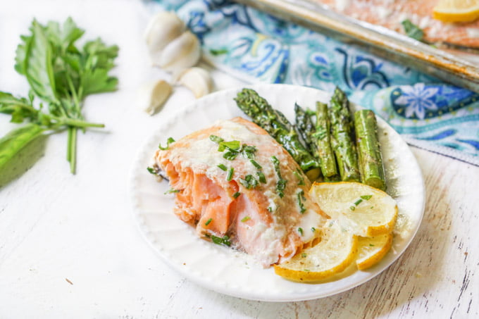 white plate with salmon, asparagus and lemon slices and fresh herbs and garlic near by