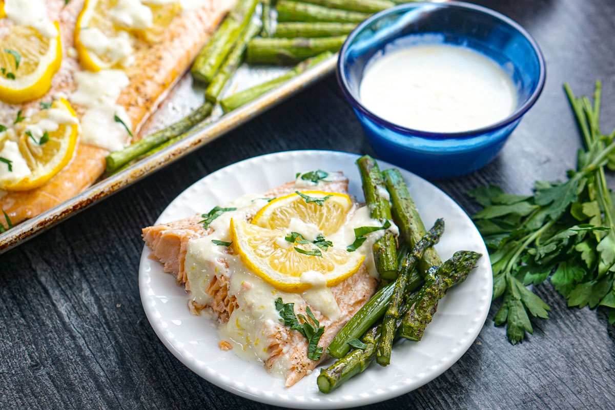 White plate with a piece of sheet pan salmon dinner and asparagus with a blue bowl of sauce in background.