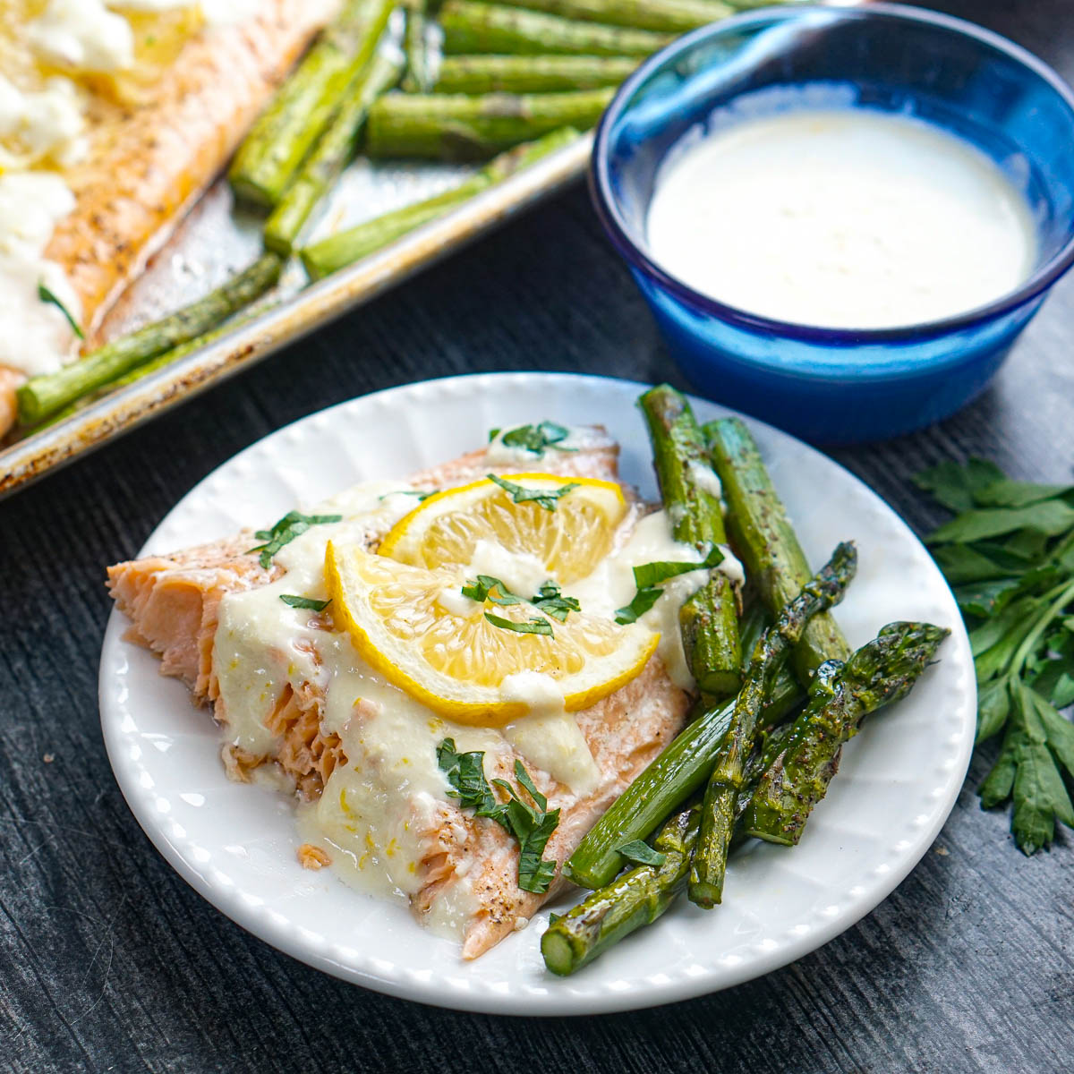 White plate with a piece of sheet pan salmon dinner and asparagus with a blue bowl of sauce in background.