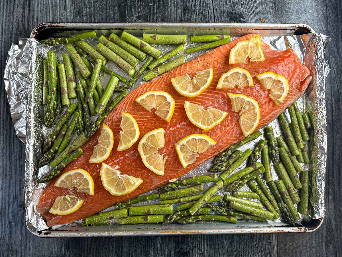 Baking sheet with raw filet covered in lemon slices and scattered asparagus.