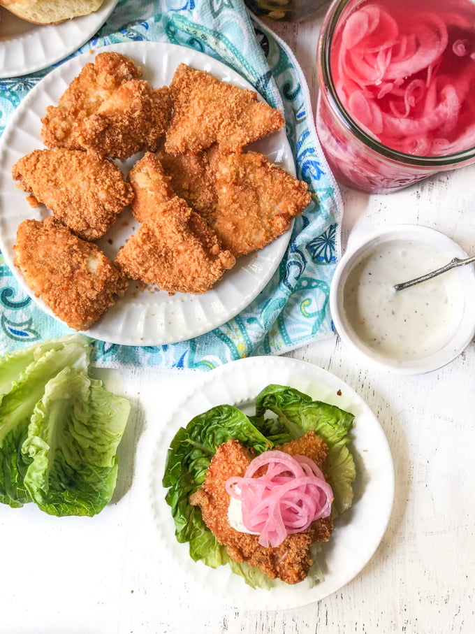 keto fried chicken sandwich using lettuce wrap on white plate and a plate of fried chicken patties and red pickled onions in a jar