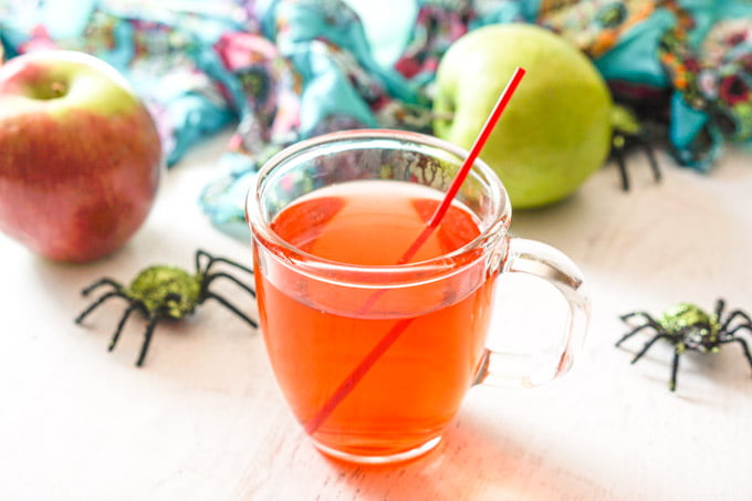 a mug of hot low carb candy apple drink with apples and decorative spiders in background