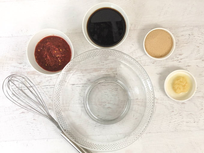 empty glass bowl, whisk and small bowls with ginger, garlic chili sauce, soy sauce and brown sugar