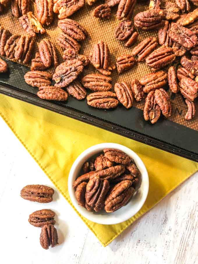 white cup with roasted pecans on yellow place mat with cookie tray in background