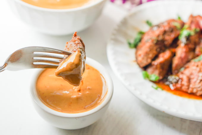 fork dipping a piece flank steak in the a little white bowl of keto peanut butter dipping sauce