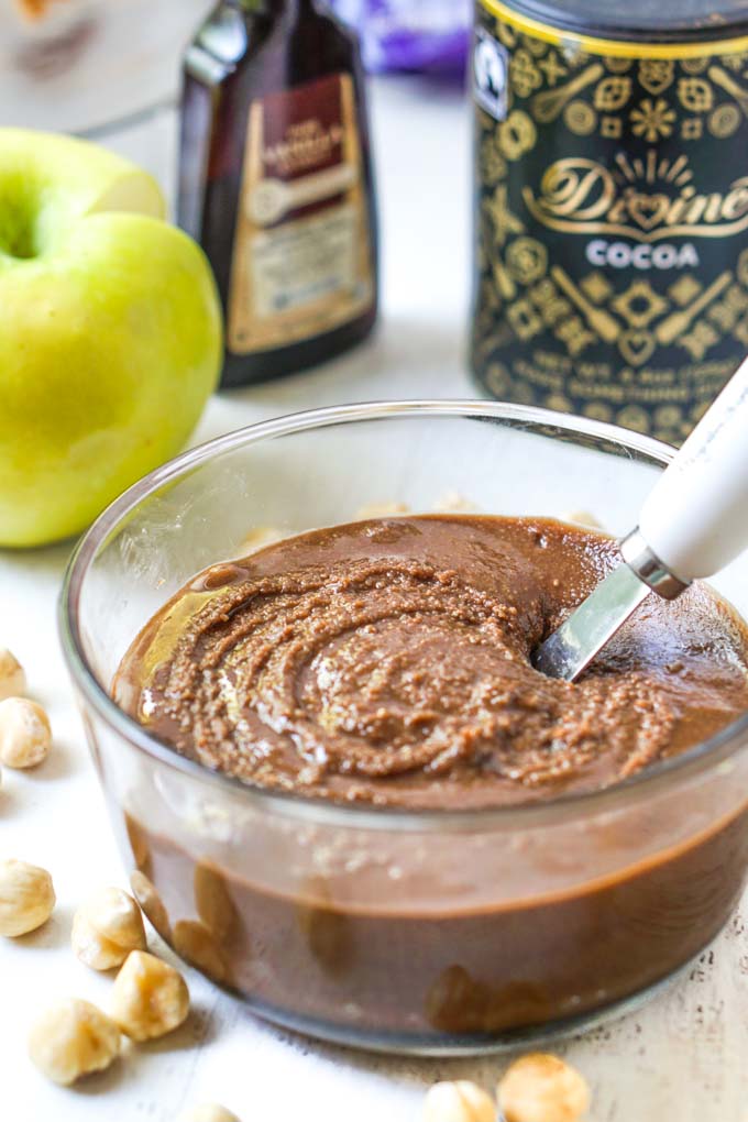 glass bowl with keto chocolate hazelnut butter with spreader knife and green apple, vanilla and cocoa in background