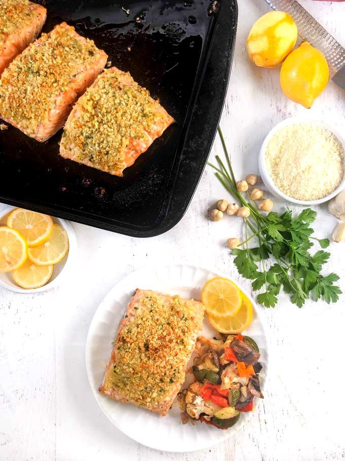 baked nut crusted salmon on cookie sheet and white plate with sliced lemons, parmesan cheese, fresh parsley and roasted hazelnuts near by