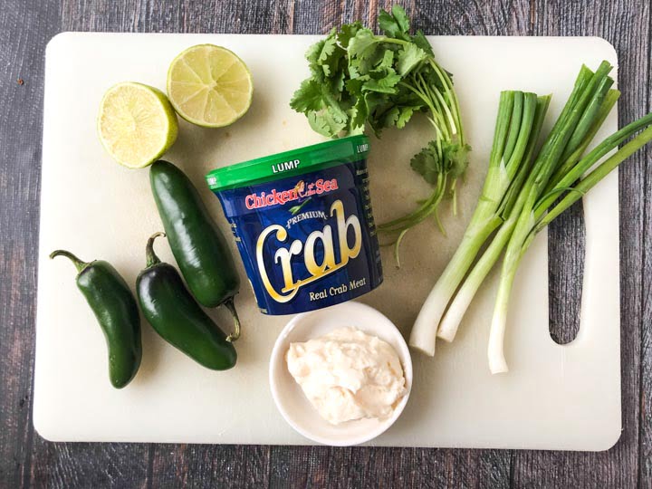 white cutting board with ingredients for easy crab cakes: sliced lime, japalenos, cilantro, green onions. bowl of mayo and lump crabmeat.