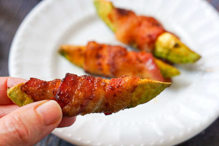 fingers holding a bacon wrapped avocado fry with the white plate in the background