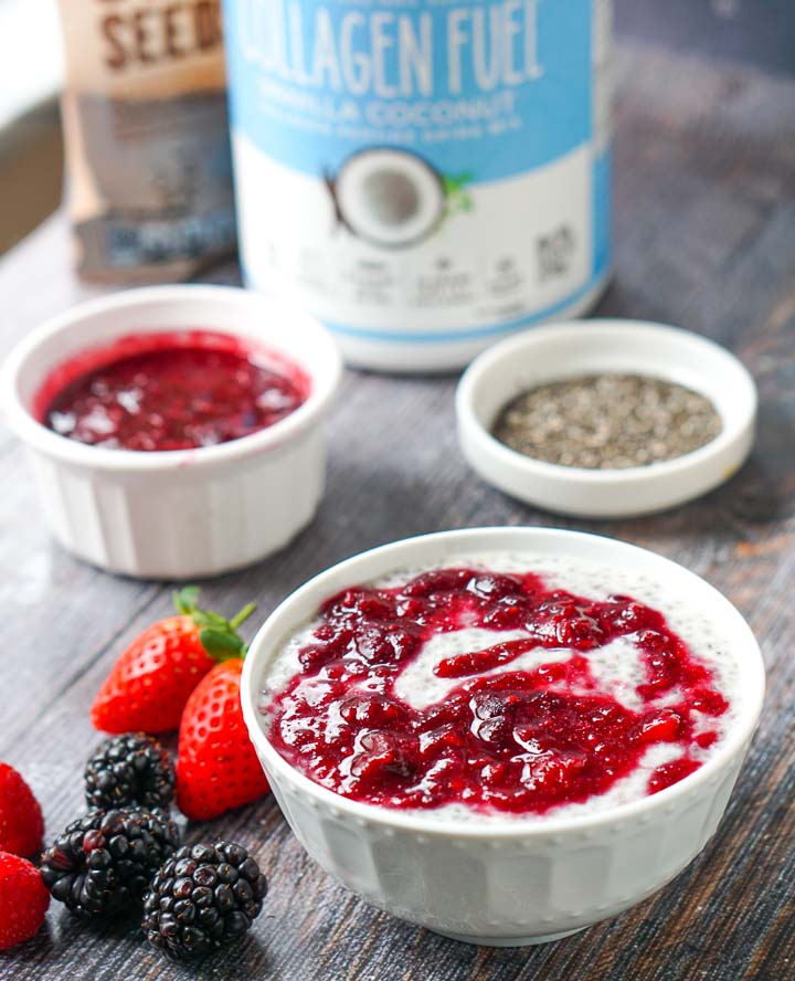 white bowl of low carb berry chia pudding with berry sauce, chia seeds and collagen fuel in background and fresh berries