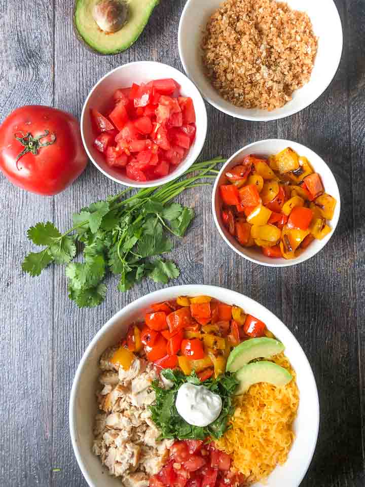white low carb chicken burrito bowl with fresh sprigs of cilantro, bowls of tomatoes and roasted peppers and half of an avocado