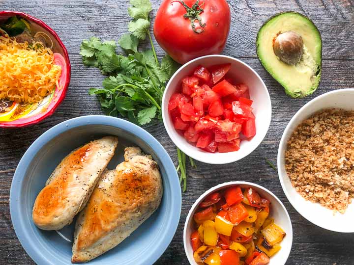 cooked ingredients for a healthy burrito bowl: chicken breasts, shredded cheddar, roasted peppers, cauliflower rice, sprigs of cilantro, tomatoes and avocado