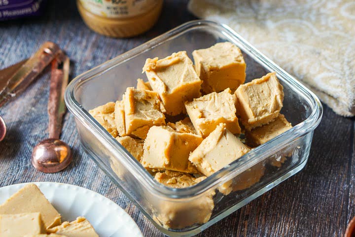 square glass container with peanut butter fudge pieces with copper measuring spoons and. tea towel in background