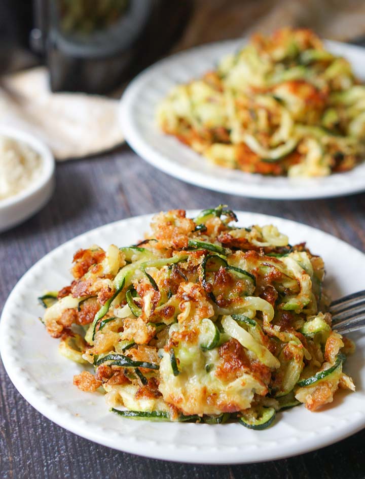 closeup of white plate with cheesy zucchini noodles and a plate in background