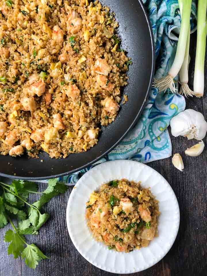 aerial view of a pan of shrimp fried cauliflower rice and a white dish with cloves of garlic, cilantro and green onions