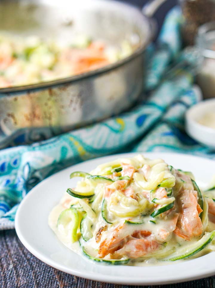 closeup of a white plate with zucchini noodles with salmon and keto alfredo sauce with pan in background
