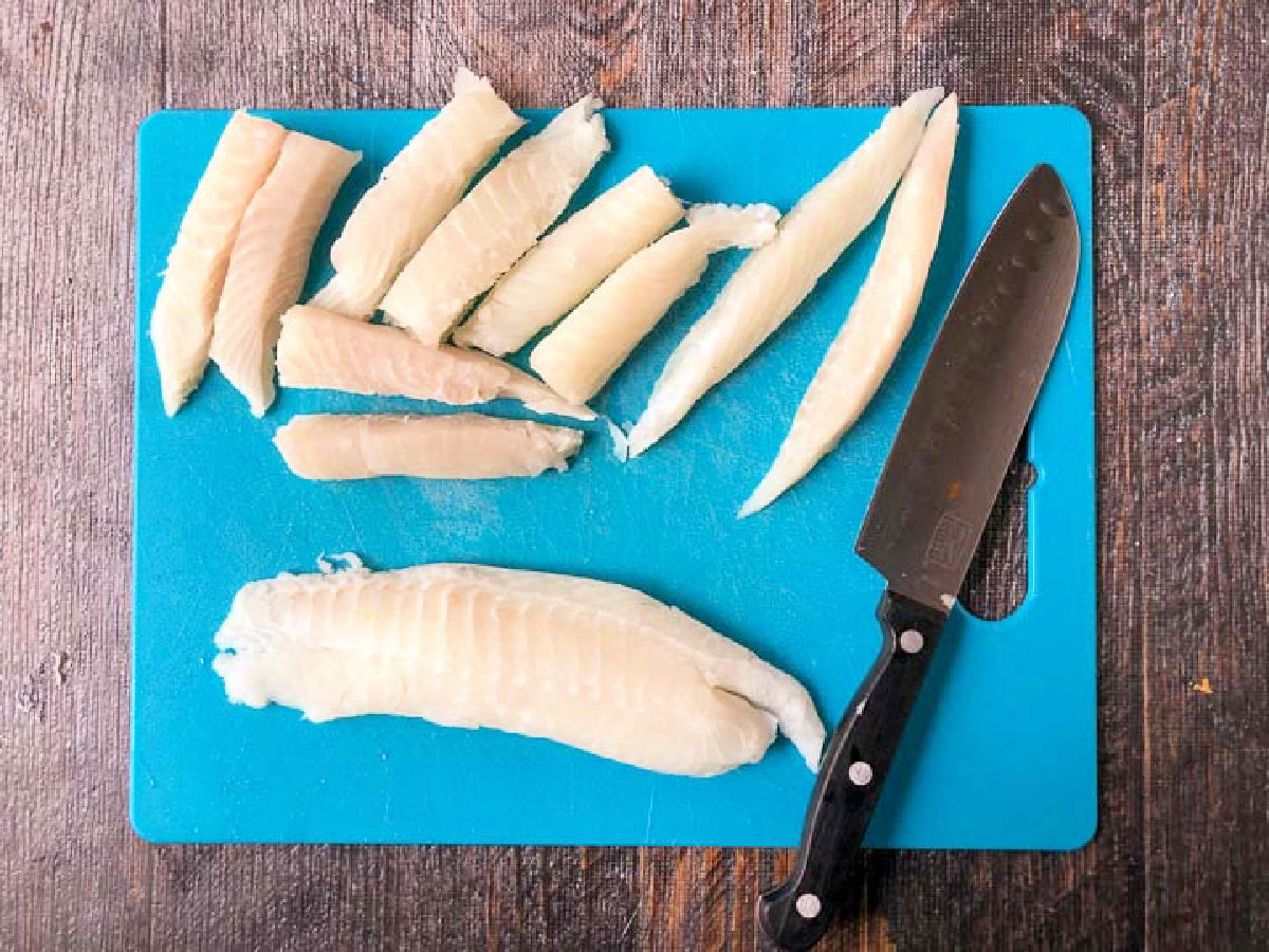 Blue cutting board with raw tilapia loins cut into sticks.