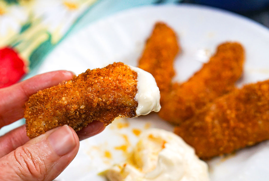 Fingers holding a fish stick dipped in tartar sauce.