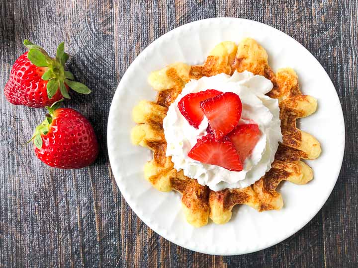 closeup of a keto chaffle with whipped cream and sliced strawberries on top and 2 fresh strawberries next to it