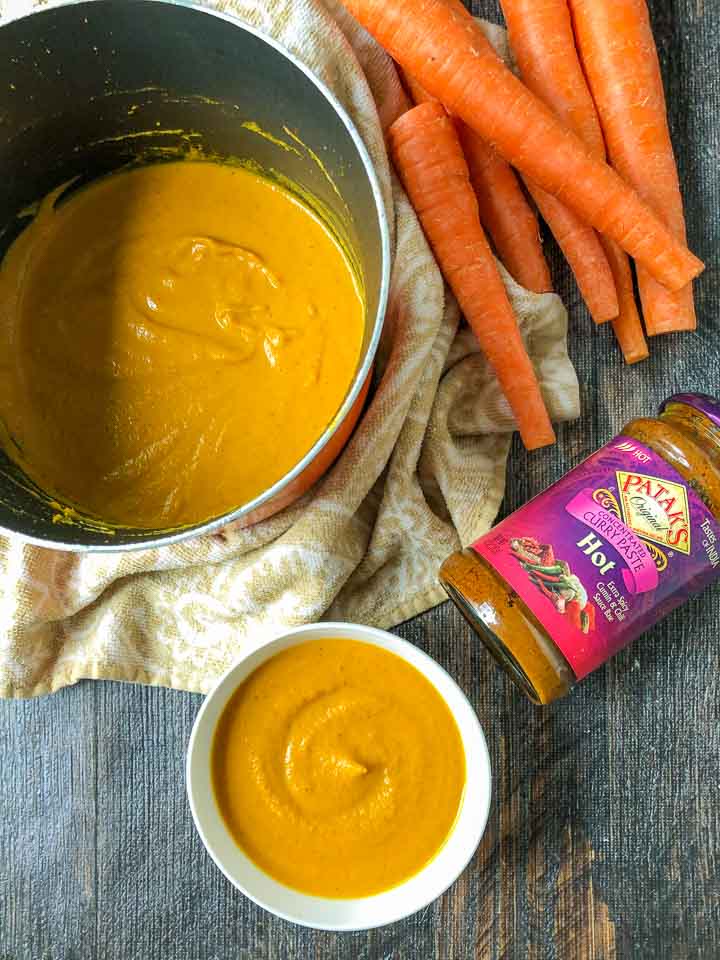white bowl and a pot of low carb creamy curry carrot soup with raw carrots and Patak's hot curry paste