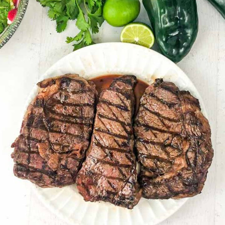 grilled steaks on a white plate with limes, cilantro and poblanos at the top of the plate