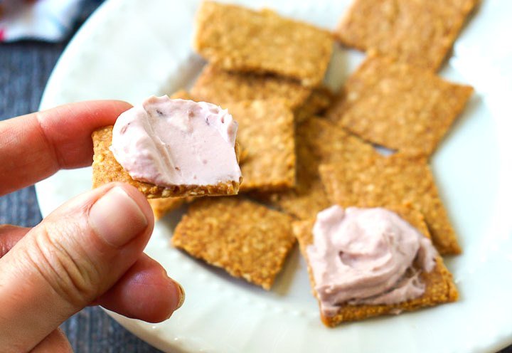 fingers holding a peanut cracker with berry cream cheese on top and a white plate in background