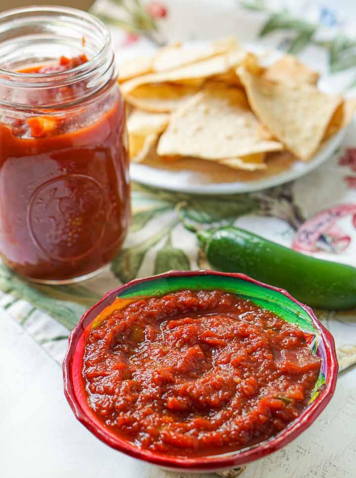 red bowl with thick homemade low carb salsa with a major jar in the background with a plate of chips and a raw jalapeno