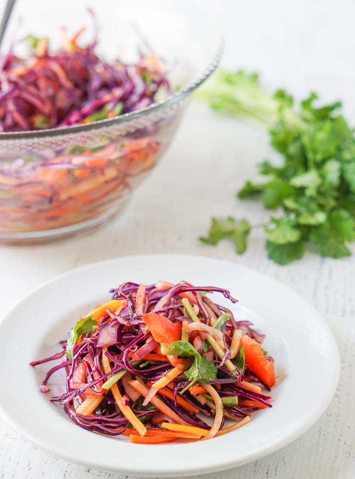 closeup of a white plate with low carb rainbow Asian slaw with bowl in background and bunch of cilantro