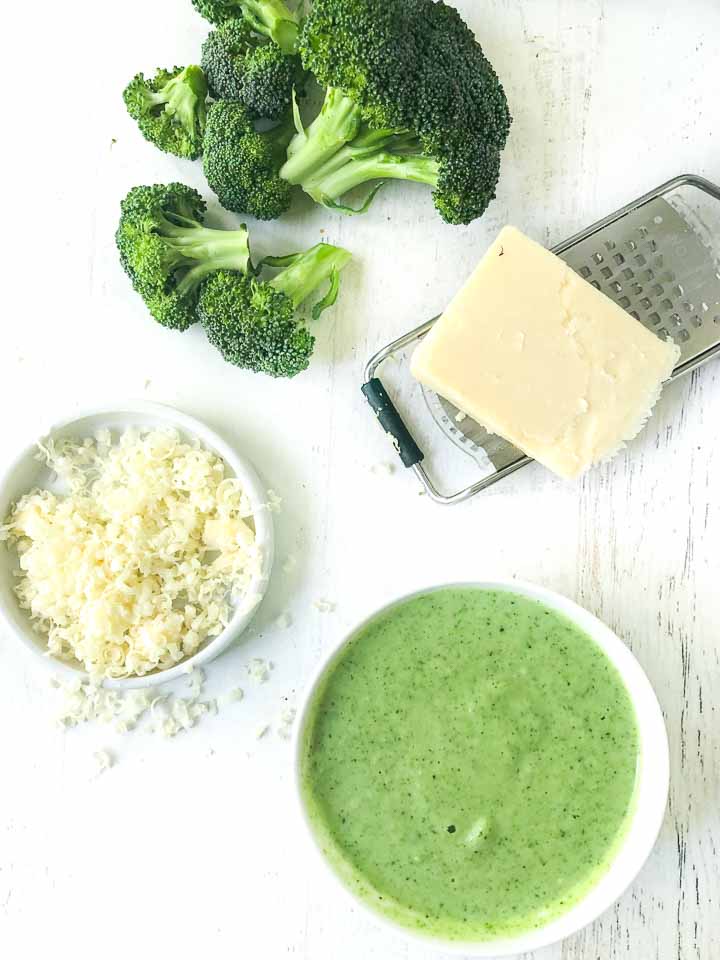bowl of broccoli soup, bowl of grated cheddar, fresh broccoli florets and a piece of cheese with a grater