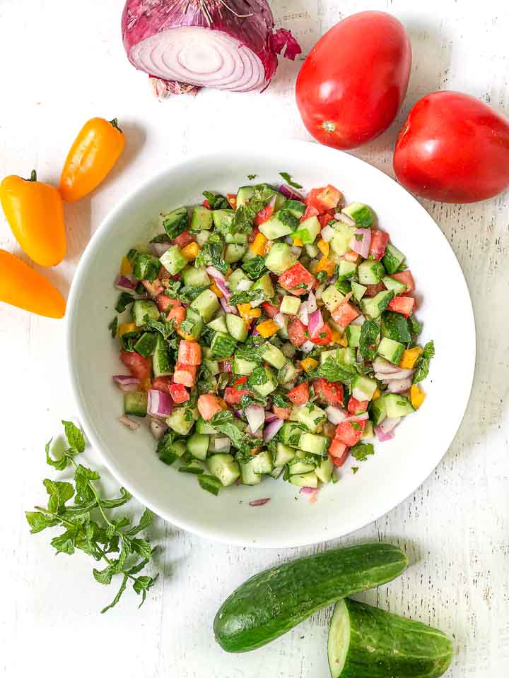aerial view of white bowl with chopped garden salad and fresh peppers, cucumber, red onion and mint around the bowl 