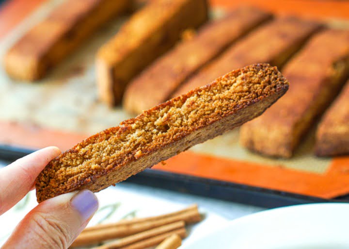 fingers holding a piece of the finished biscotti with a trayful of it in the background