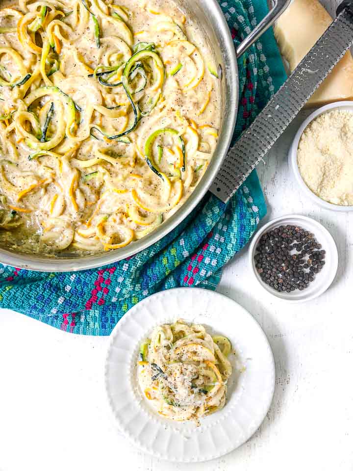 pan and white plate with cheesy zucchini noodles with microplane and block of cheese and black peppercorns