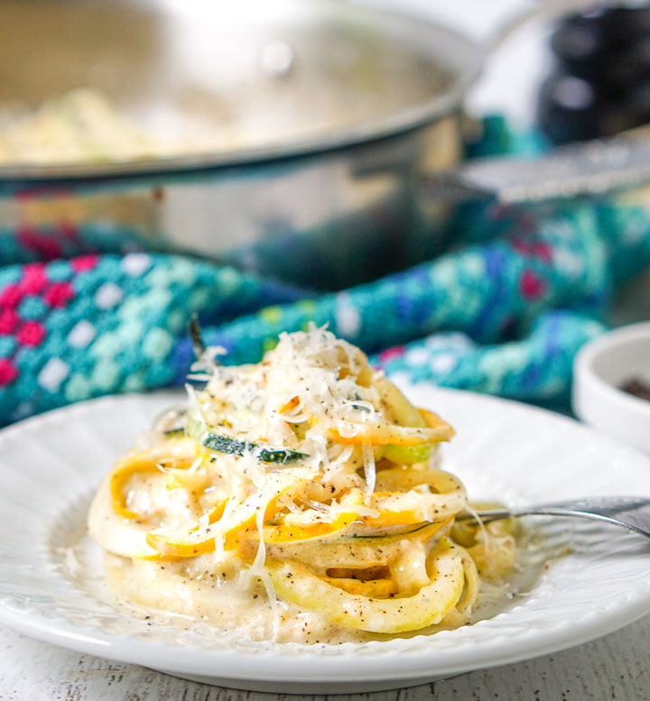 closeup of a plate of cheesy zoodles with grated Parmesan on top and a pan and bright blue tea towel in background