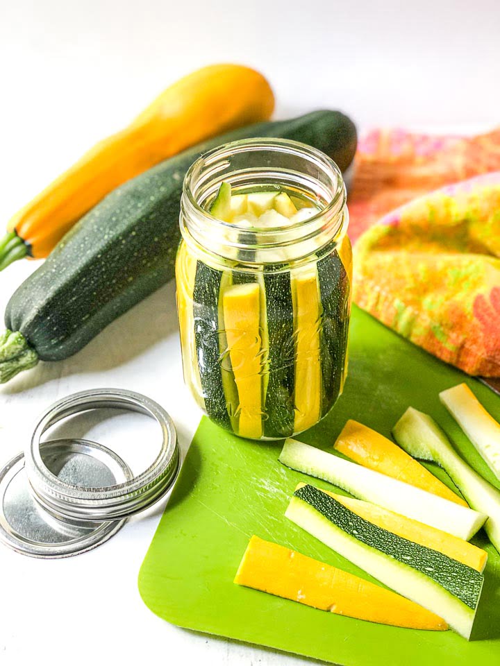 jar with yellow and green pickled zucchini on a green cutting board with raw zucchini chopped, and silver lids