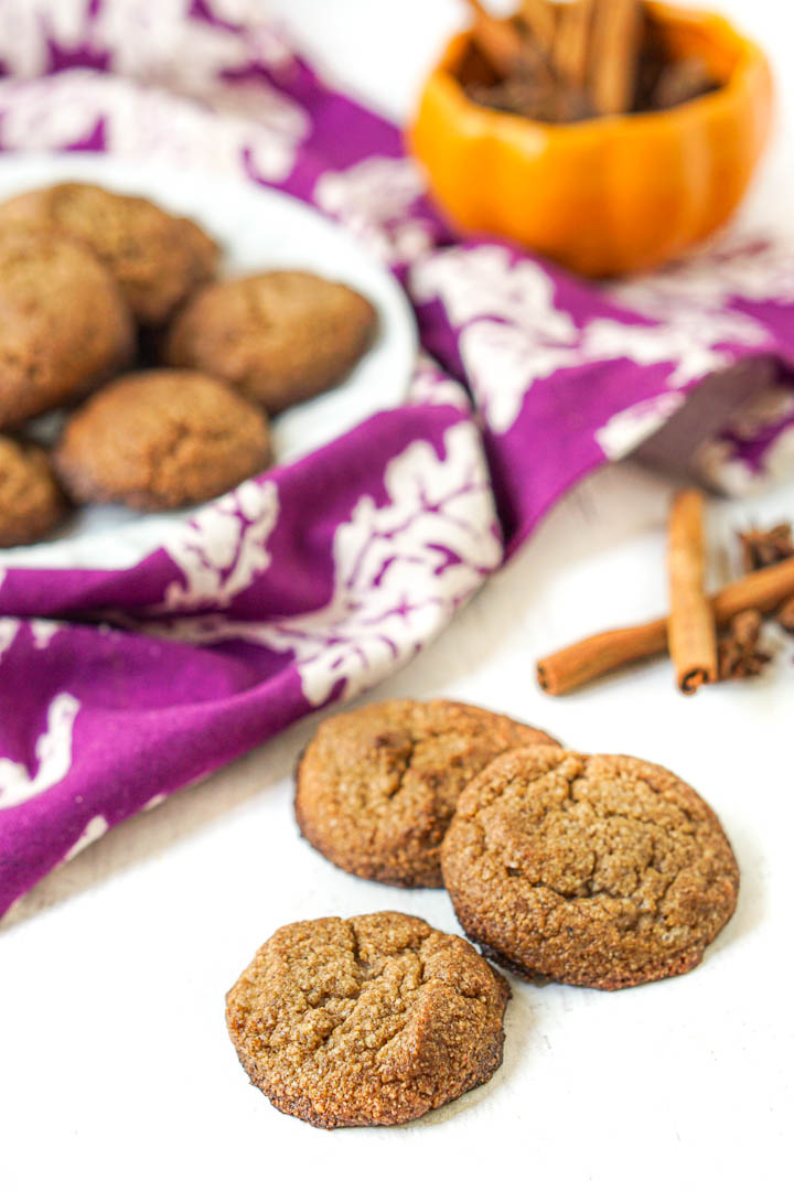 3 gluten free cookies with orange pumpkin, white plate of cookies and purple tea towel in background