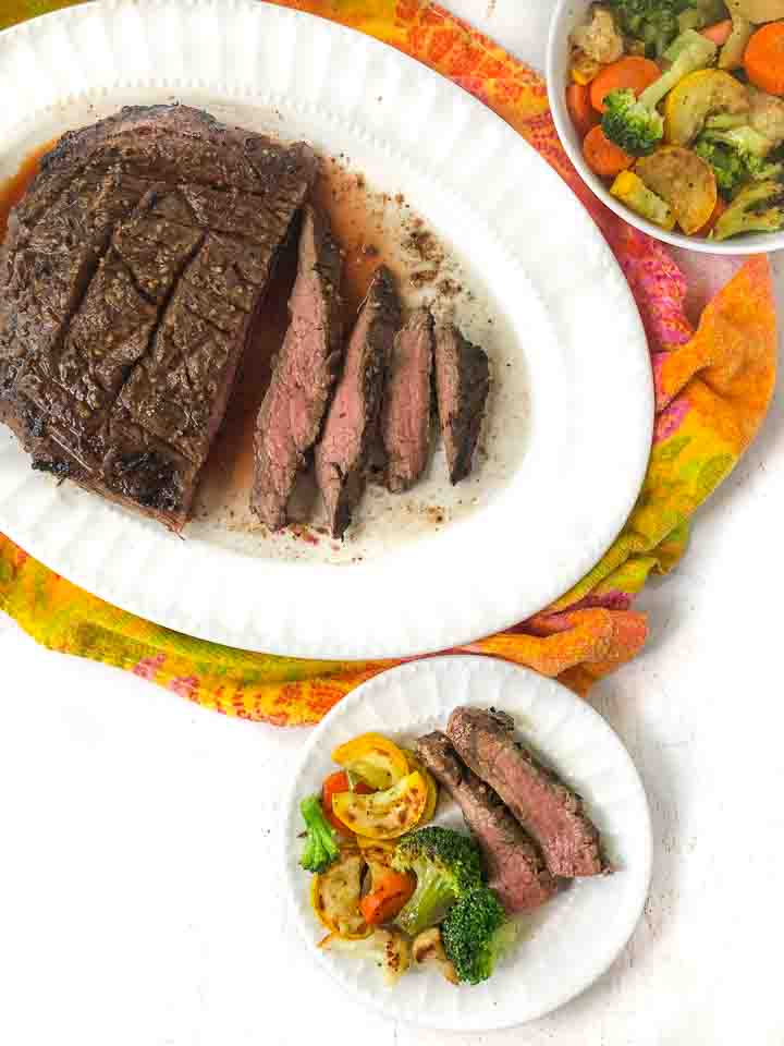 white plater and plate with grilled flank steak and a bowl of roasted veggies on a colorful tea towel