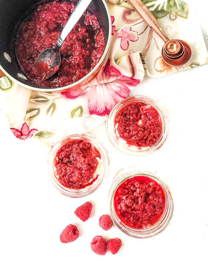 3 keto cheesecake jars with pan of sugar free raspberry sauce and fresh scattered raspberries
